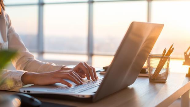 A businesswoman types on a laptop as the sun sets through the large windows behind her